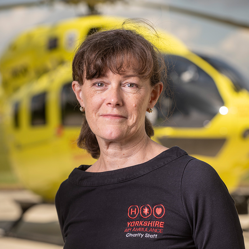 A lady with dark hair, wearing a black top is standing in front of a yellow helicopter