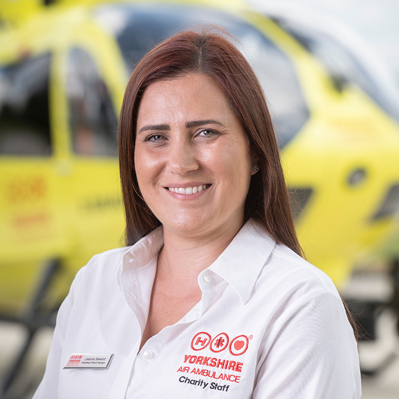 A lady with long dark hair, wearing a white shirt, stands in front of a yellow helicopter