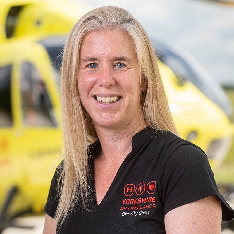 A lady with long blonde hair, wearing a black top is standing in front of a yellow helicopter