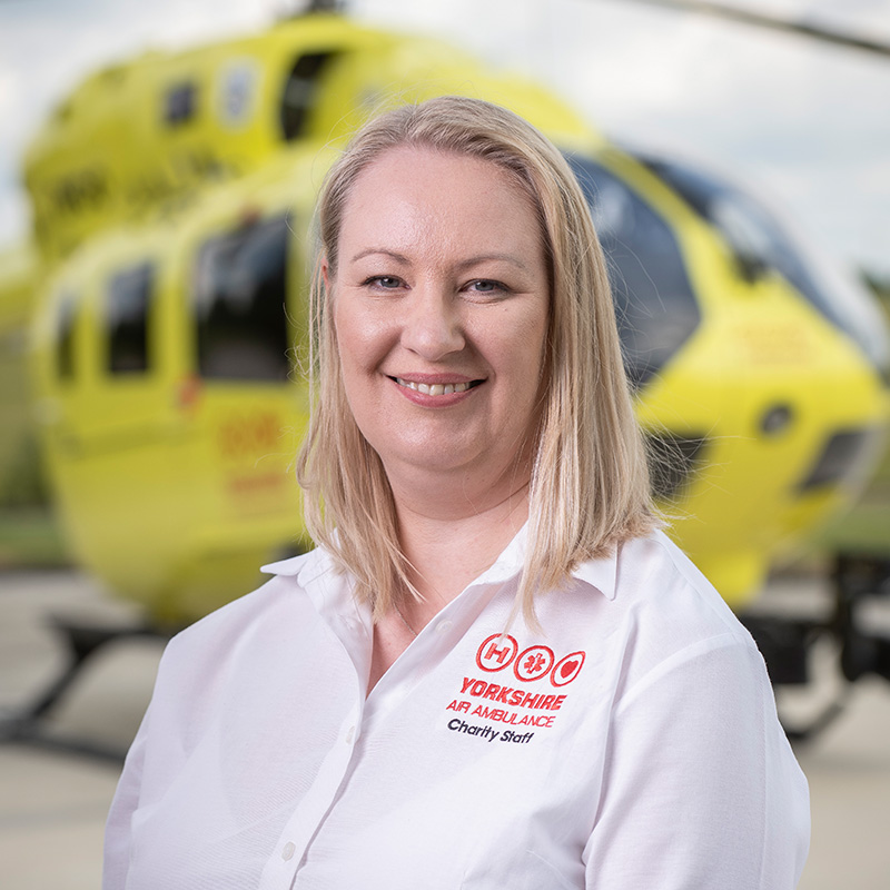 A lady with shoulder length blonde hair, wearing a white shirt is standing in front of a yellow helicopter