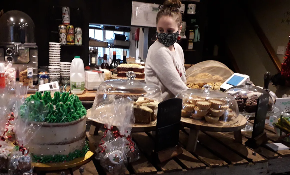 Cakes and buns on a counter in a tea room. A lady is behind the counter. She is wearing a white jumper and a black and white face covering. She has brown hair which is tied up in a bun.