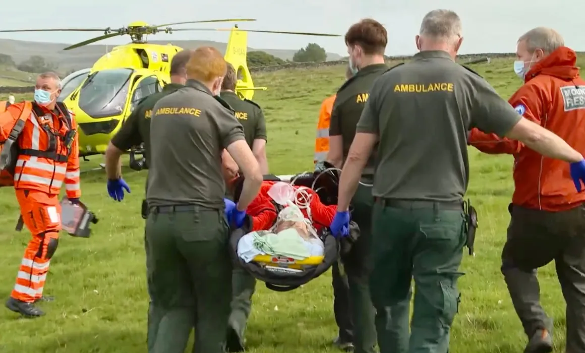 Picture shows Ambulance Paramedics, Yorkshire Air Ambulance crew and Mountain Rescue Team Members carrying a lady on a stretcher towards the yellow Yorkshire Air Ambulance helicopter across a grass field in the countryside