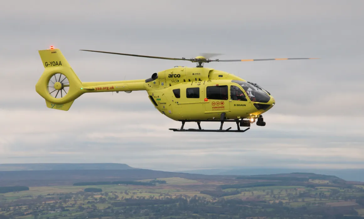 A yellow helicopter flying over fields and hills.