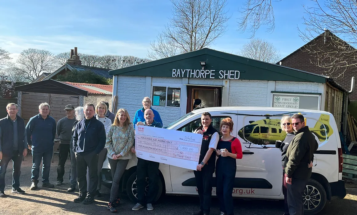 A group of people are stood in front of a people with a sign which reads 'BAYTHORPE SHED'. Two of them are holding a large cheque, which is made payable to Yorkshire Air Ambulance and is for £1000. One of those people is wearing a Yorkshire Air Ambulance gilet and they are stood in front of a white van which has a yellow Yorkshire Air Ambulance helicopter on it. There is also a garage to the left of the image behind the group of people and the roofs of houses are visible beyond that. There are a number of trees also visible in the background.