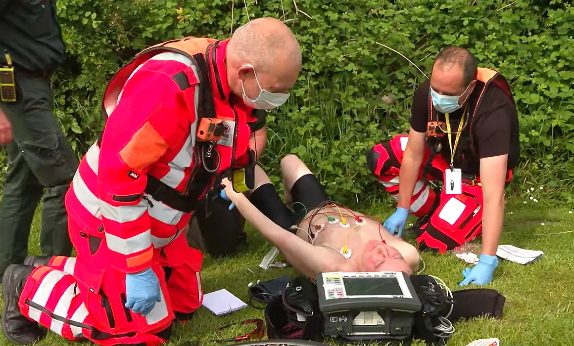 Two medical personnel are tending to a gentleman who is laying on the grass. There is medical equipment around them. The two medical personnel are wearing masks. One is wearing an orange jacket and trousers, the other orange trousers and a black t-shirt. They are knelt beside the man.