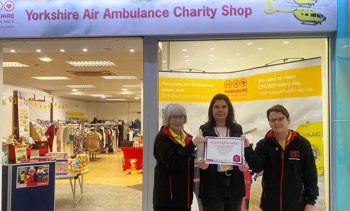 Two Yorkshire Air Ambulance volunteers and fundraiser Clare Deacon, are holding a volunteer award. They are stood outside the Yorkshire Air Ambulance charity shop.