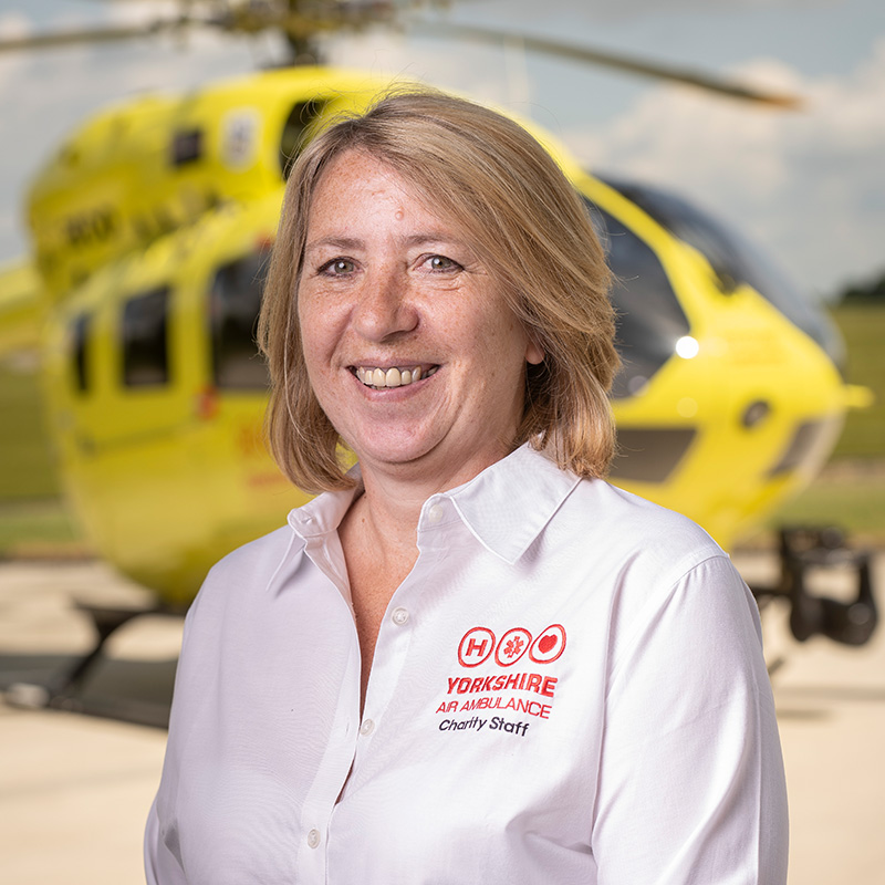 A women with light coloured, shoulder length blonde hair, wearing a white shirt is standing in front of a yellow helicopter