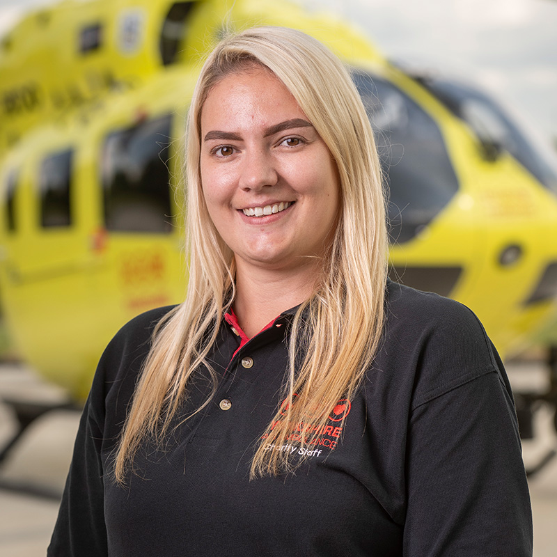 A lady with long blonde hair, wearing a black top is standing in front of a yellow helicopter
