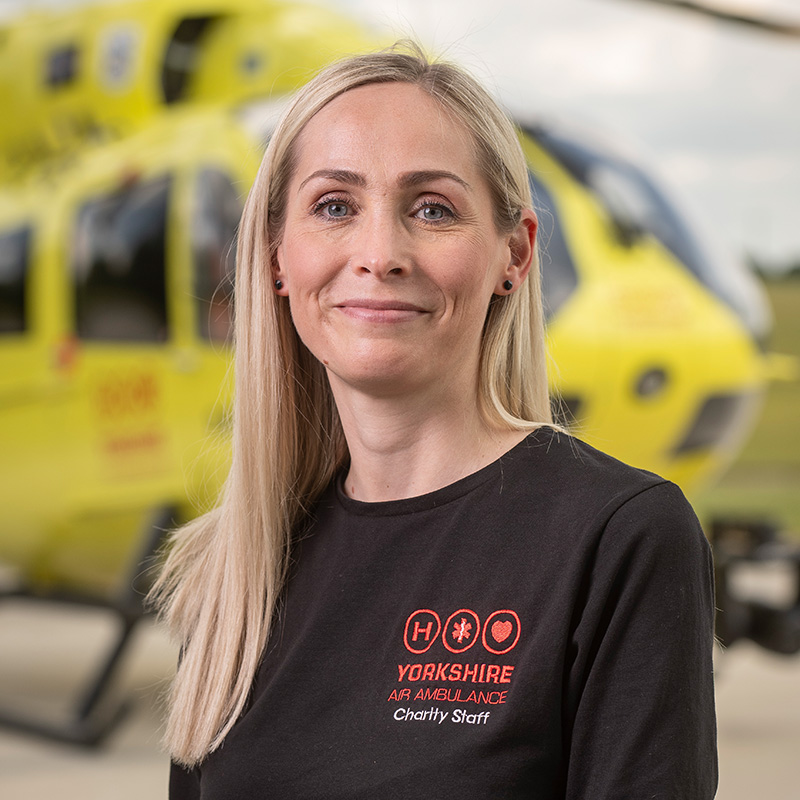 A lady with long blonde hair, wearing a black top is standing in front of a yellow helicopter