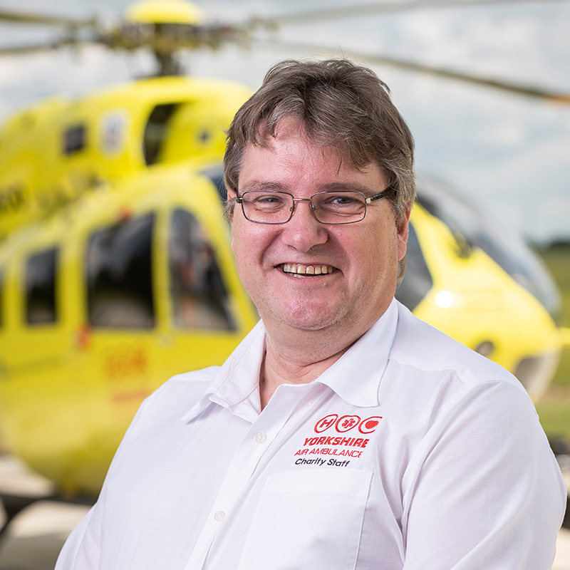 A gentleman with dark hair and glasses, wearing a white shirt, stands in front of a yellow helicopter