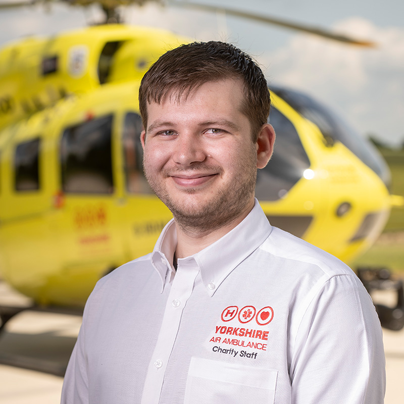 A man with short dark hair, wearing a white shirt is standing in front of a yellow helicopter