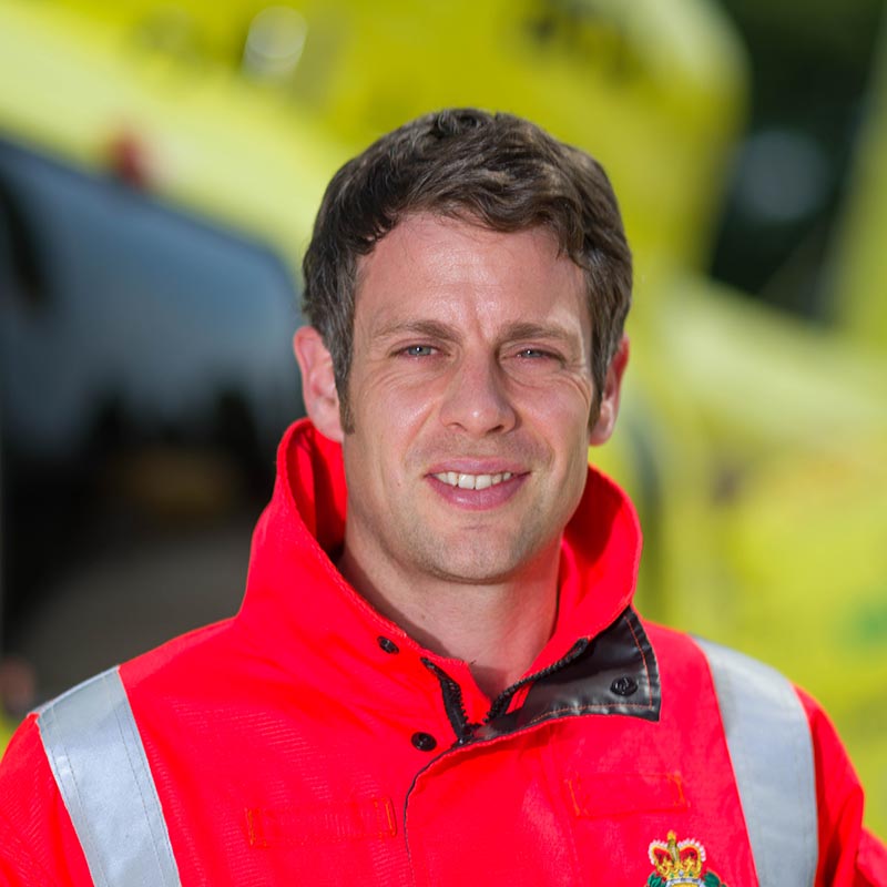 A man with medium brown hair wearing an orange jacket stands in front of a yellow helicopter