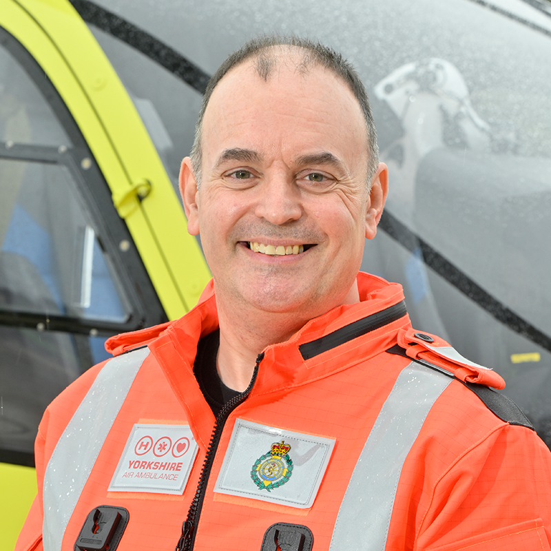 A man wearing an orange flight suit, standing in front of a yellow helicopter.