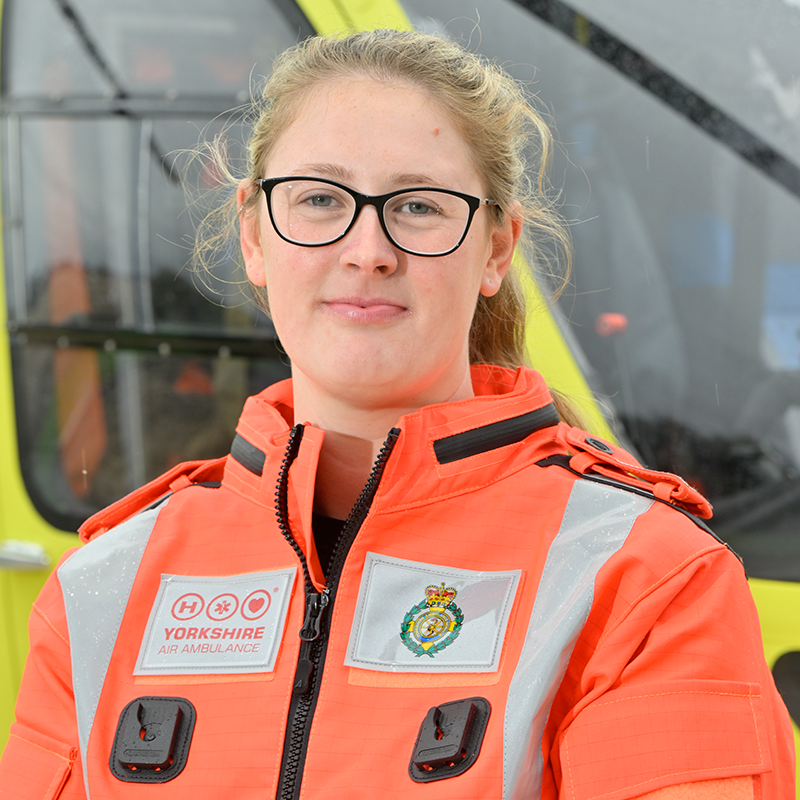 A woman wearing glasses and an orange flight suit, standing in front of a yellow helicopter.