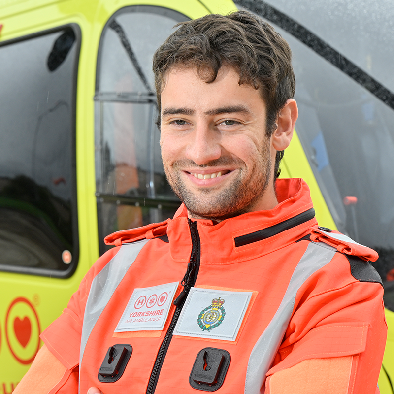 A man wearing an orange flight suit, standing in front of a yellow helicopter.