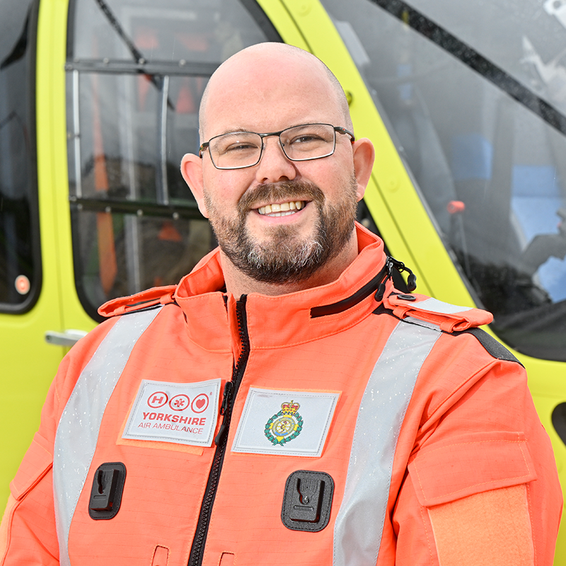 A man with a beard, wearing glasses and an orange flight suit, standing in front of a yellow helicopter.