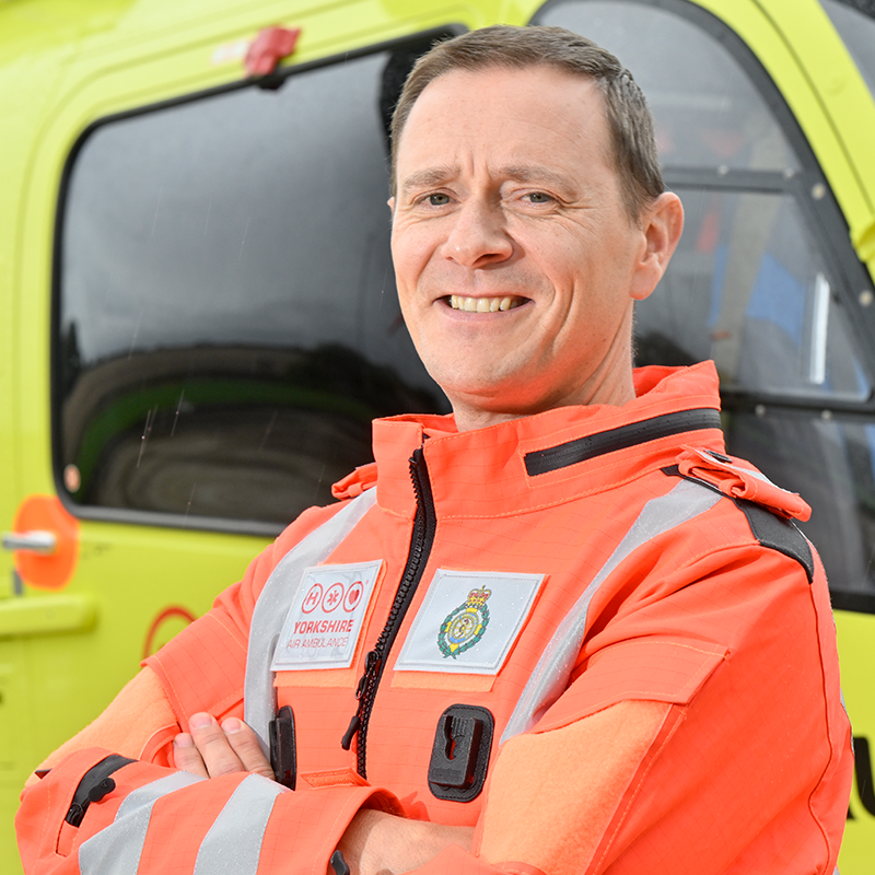 A man wearing an orange flight suit, standing in front of a yellow helicopter.