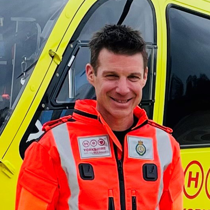 Man with short brown hair wearing an orange flight suit standing in front of a yellow helicopter.