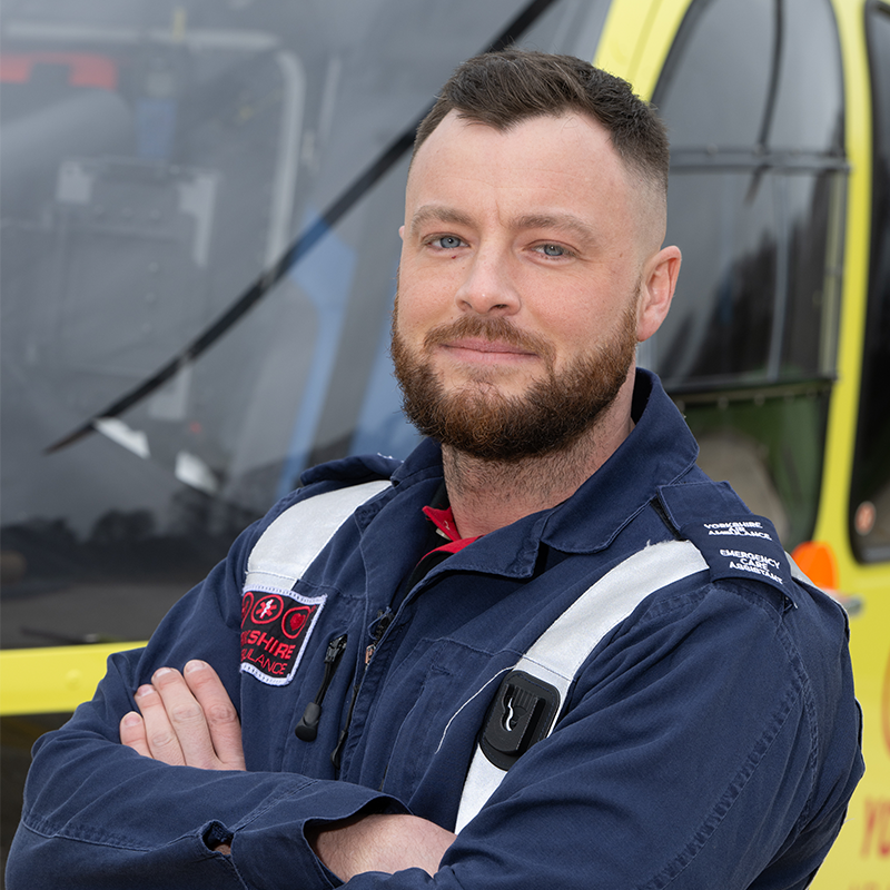 A man with dark hair a beard wearing a blue flight suit, stood with his arms crossed in front of a yellow helicopter.